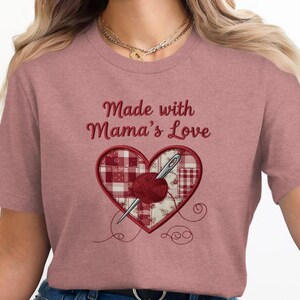 May include: A dusty pink t-shirt with the embroidered phrase "Made with Mama's Love." The design features a heart shape with a needle, yarn, and a red and white patchwork pattern. Decorative stitching is included in the design.
