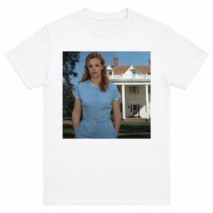 May include: White t-shirt featuring a still from the movie "The Notebook." The image shows a woman in a light blue dress standing in front of a white house with columns. The woman has red hair and is looking at the camera.