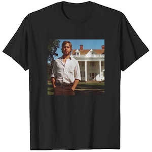 May include: Black t-shirt featuring a color image of a man in a white shirt and brown pants standing in front of a white house with a blue sky background. The man has his hands in his pockets.