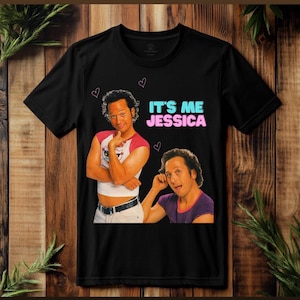May include: Black t-shirt featuring a graphic of a man in two poses, with the text "IT'S ME JESSICA" in blue and pink. The design includes small pink hearts. The shirt is displayed on a wooden surface.