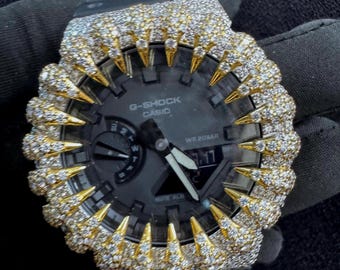 May include: A G-Shock Casio watch with a black face and a bezel encrusted with sparkling crystals and gold-colored accents. The watch features a black band and the text "G-SHOCK CASIO" on the face. The watch is water resistant to 20 bar.