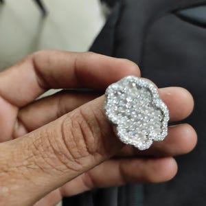 Pode incluir: Um anel prateado com um design floral, incrustado com numerosas pedras preciosas pequenas e transparentes. O anel é segurado por uma mão, mostrando os detalhes intrincados da joia. As pedras preciosas brilham.