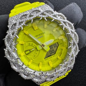 Op de afbeelding: Een neongele G-Shock Casio-horloge met een met diamanten bezette lunette. De wijzerplaat van het horloge is ook geel, met een digitale weergave van de tijd. De horlogeband is ook neongeel.