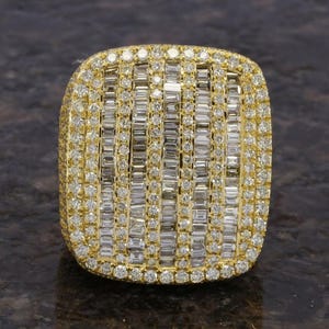 Pode incluir: Um grande anel de ouro retangular, cravejado com numerosos diamantes. O anel apresenta um design central de diamantes de corte baguette verticais, rodeado por uma borda de diamantes redondos menores. Fundo escuro.