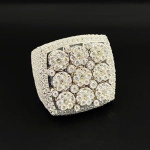 Pode incluir: Um anel prateado de rosto quadrado, incrustado com numerosas pedras preciosas claras e brilhantes. A face do anel apresenta um grupo de pedras maiores rodeadas por outras menores, criando uma exibição deslumbrante. A banda também é coberta por pedras preciosas menores.