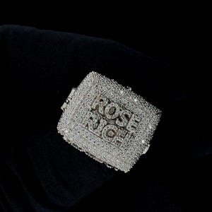 Pode incluir: Um anel quadrado prateado incrustado com numerosas pedras preciosas pequenas e transparentes. A superfície do anel apresenta o texto "ROSE RICH" numa fonte estilizada. O anel está sobre um fundo escuro.