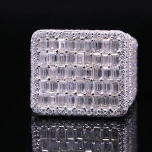 Pode incluir: Um anel prateado de formato quadrado, adornado com uma multidão de pedras preciosas retangulares e transparentes. A superfície principal é densamente adornada com estas pedras, e as bordas são bordadas com pedras menores e redondas, criando uma aparência brilhante e luxuosa.