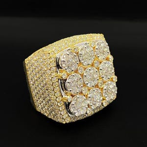 Pode incluir: Um anel grande, dourado, incrustado com numerosos diamantes pequenos e brilhantes. O anel apresenta uma parte superior retangular com um grupo de diamantes maiores no centro, rodeados por pedras menores. A banda e as laterais também são cobertas por diamantes.