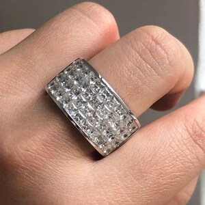 Pode incluir: Um anel retangular de prata com uma superfície coberta por pequenas pedras preciosas claras, cortadas em forma quadrada. O anel está em um dedo, mostrando os detalhes intrincados do design e o brilho das pedras.