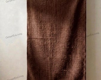 May include: A brown, rectangular rug hanging on a rod. The rug has a textured surface and is suspended against a white wall. The rug is a solid color and appears to be made of a soft material.