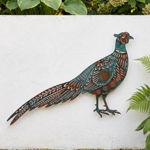 Op de afbeelding: Een metalen wanddecoratie in de vorm van een fazant, met een patina-afwerking in tinten blauwgroen, koper en bruin. De vogel heeft ingewikkelde uitgesneden patronen en geëtste details op het lichaam, de vleugels en de lange staartveren.