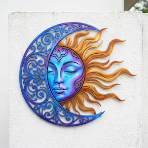 May include: A celestial wall hanging featuring a sun and moon design. The sun has a serene face with closed eyes, and the moon is a crescent shape with intricate, swirling patterns. The color palette includes blue, purple, and orange.