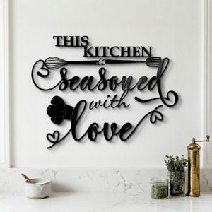 Op de afbeelding: Zwarte metalen keukenmuurkunst met de woorden "THIS KITCHEN is SEASONED with LOVE". De kunst toont een garde, vork en koksmuts. De kunst is gemonteerd op een witte muur boven een aanrecht.