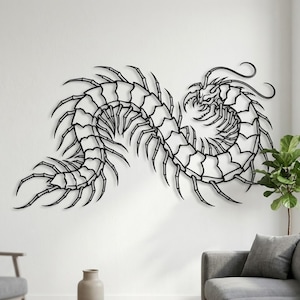May include: A black metal wall art piece depicting a centipede-like creature. The artwork is mounted on a white wall, with a sofa, a vase, and a plant in the background. The creature has many legs and a detailed head.