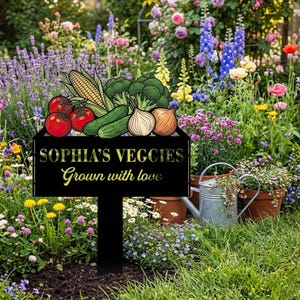 以下が含まれることがあります： 「SOPHIA'S VEGGIES Grown with love」の文字と野菜のイラストが描かれた黒い金属製のガーデンサイン。バラやラベンダーなど、色とりどりの花が咲き誇る庭に設置されています。