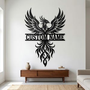 Puede incluir: Arte mural de metal negro con un fénix con las alas extendidas y el texto "CUSTOM NAME" en el centro. La pieza decorativa está montada en una pared blanca sobre un mueble de madera.