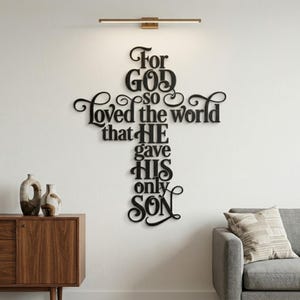 May include: Black wooden cross-shaped wall art with the text "For God so loved the world that He gave His only Son." The art is mounted on a white wall, with a modern light fixture above. A wooden cabinet and gray sofa are in the background.