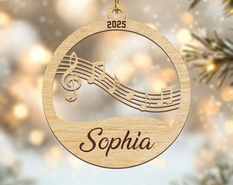 Pode incluir: Enfeite de Natal de madeira com tema musical. O enfeite redondo apresenta um design vazado com notas musicais e uma clave de sol. O nome "Sophia" está gravado na parte inferior e o ano "2025" na parte superior.