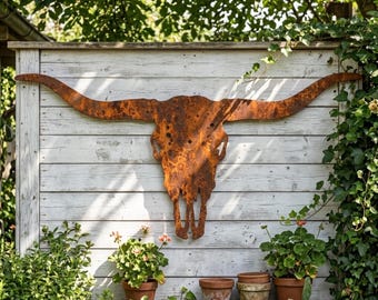 May include: A large, rusty metal longhorn skull mounted on a white wooden fence. The skull has long, curved horns and is surrounded by greenery and potted plants, creating a rustic, outdoor aesthetic.