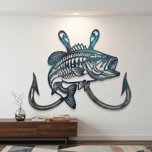 Pode incluir: Uma obra de arte de parede em metal que retrata um peixe azul esverdeado com dois grandes ganchos. O peixe tem detalhes recortados. Os ganchos são de cor cinza escuro. A obra está montada numa parede branca.