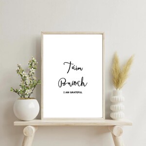 May include: A framed print with the words "Táim Buíoch" and "I AM GRATEFUL" in black script. The print is displayed on a light wooden shelf with white vases and floral arrangements on either side.
