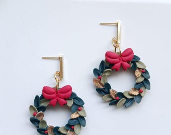 May include: A pair of dangle earrings featuring circular wreaths made of dark green, light green, and gold glitter leaves. Each wreath is accented with a small red bow at the top and tiny red berries, suspended from a gold-toned metal bar post.