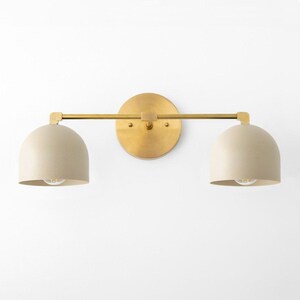 May include: A two-light vanity light fixture with a brass finish. The fixture has a circular backplate, a horizontal bar, and two beige, dome-shaped shades. Each shade houses a light bulb. The fixture is designed for wall mounting.