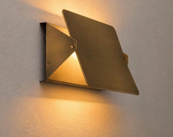 May include: A modern, geometric wall sconce made of brass. The fixture has a square base with a triangular light opening and a hinged, rectangular shade. The sconce is mounted on a textured, off-white wall, with warm light emanating from the fixture.
