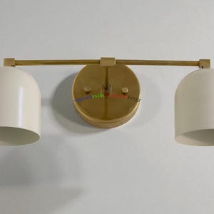 May include: A modern wall-mounted light fixture with two cream-colored, dome-shaped shades. The fixture has a gold-toned, circular base and a horizontal bar connecting the shades. The design is minimalist and stylish.