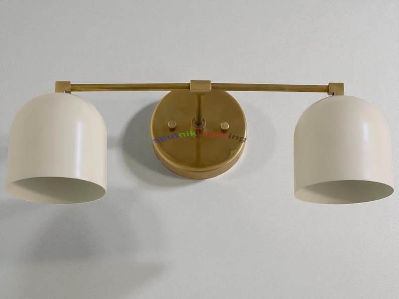 May include: A modern wall-mounted light fixture with two cream-colored, dome-shaped shades. The fixture has a gold-toned, circular base and a horizontal bar connecting the shades. The design is minimalist and stylish.