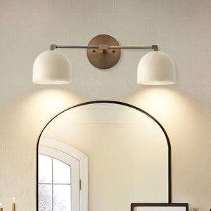 May include: A two-light vanity fixture with a bronze finish. The fixture has two white, dome-shaped shades and is mounted above a black-framed arched mirror. The wall is a light beige color.