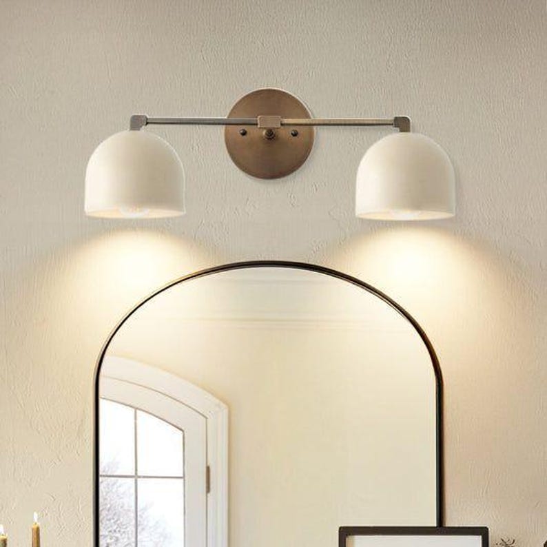 May include: A two-light vanity fixture with a bronze finish. The fixture has two white, dome-shaped shades and is mounted above a black-framed arched mirror. The wall is a light beige color.