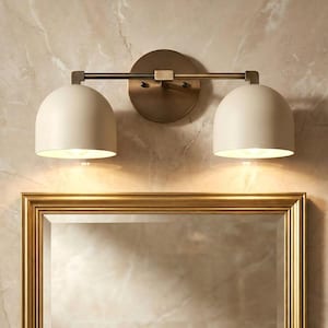 May include: A two-light vanity light fixture with a brass finish. The fixture has two off-white, dome-shaped shades. The light is mounted above a gold-framed mirror, against a marble-patterned wall.