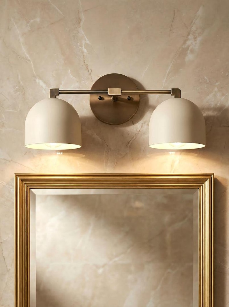 May include: A two-light vanity light fixture with a brass finish. The fixture has two off-white, dome-shaped shades. The light is mounted above a gold-framed mirror, against a marble-patterned wall.