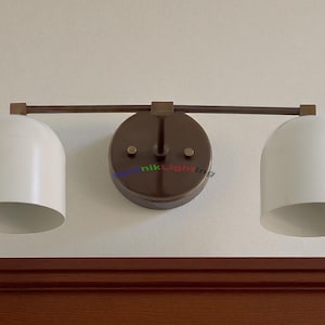 May include: A two-light wall sconce with a dark bronze finish. The fixture features two white, dome-shaped shades and a circular backplate. The sconce is mounted on a light-colored wall above a dark wood trim.