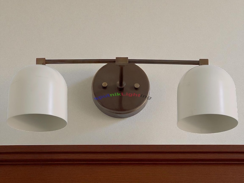 May include: A two-light wall sconce with a dark bronze finish. The fixture features two white, dome-shaped shades and a circular backplate. The sconce is mounted on a light-colored wall above a dark wood trim.