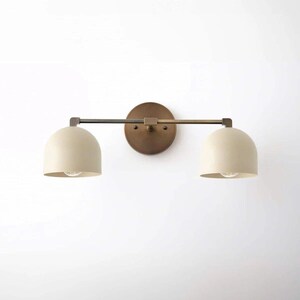 May include: A two-light vanity light fixture with a round, bronze-colored backplate. Two off-white, dome-shaped shades are connected by a horizontal bar. The fixture has a minimalist, modern design, suitable for bathrooms or hallways.