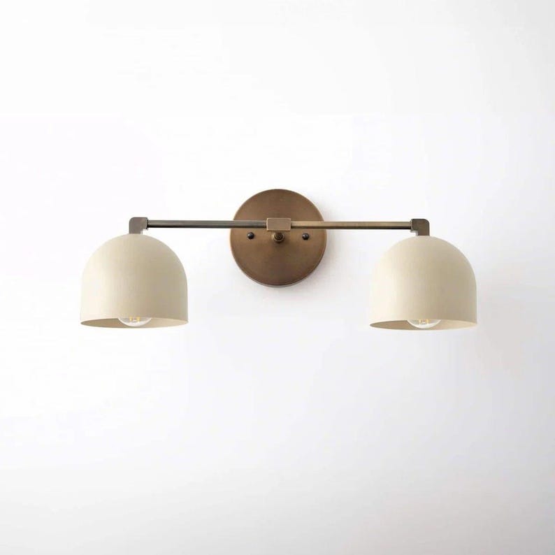 May include: A two-light vanity light fixture with a round, bronze-colored backplate. Two off-white, dome-shaped shades are connected by a horizontal bar. The fixture has a minimalist, modern design, suitable for bathrooms or hallways.