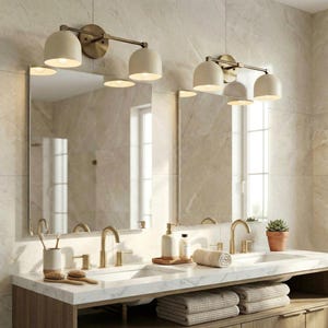 May include: Bathroom vanity with two sinks, mirrors, and lighting fixtures. The fixtures have a brass finish and off-white shades. The vanity has a marble countertop, gold faucets, and a wooden base. Accessories include soap dispensers, towels, and a small potted plant.
