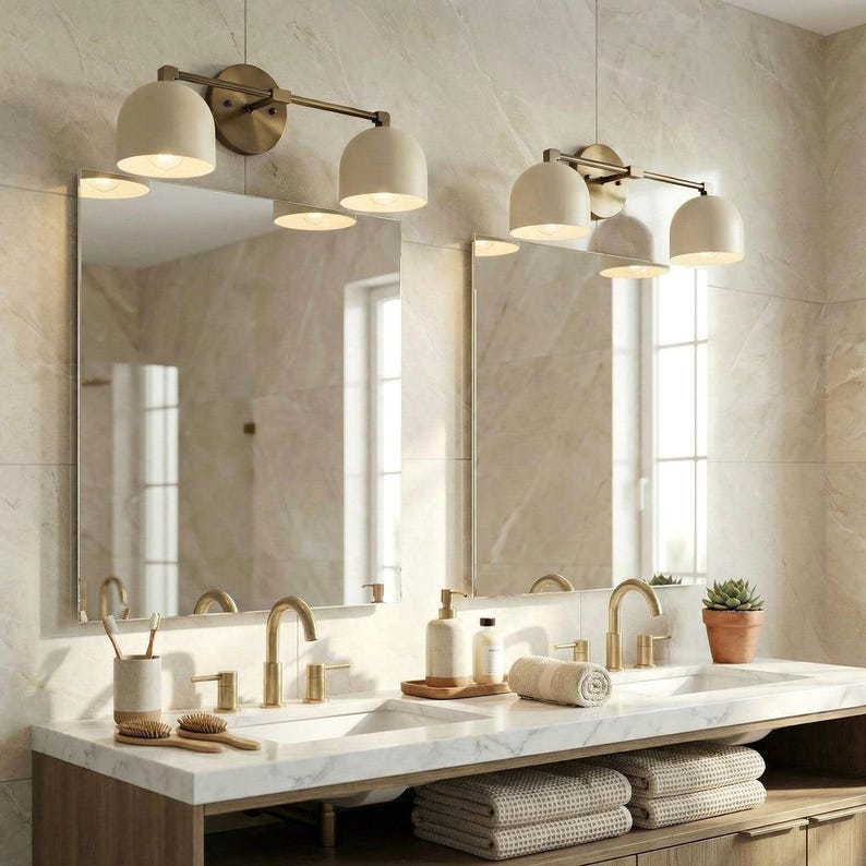 May include: Bathroom vanity with two sinks, mirrors, and lighting fixtures. The fixtures have a brass finish and off-white shades. The vanity has a marble countertop, gold faucets, and a wooden base. Accessories include soap dispensers, towels, and a small potted plant.