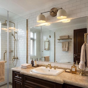 May include: A bathroom with a white tiled wall, a large mirror, and a dark brown vanity. The vanity has a marble countertop with a white sink and gold fixtures. A gold light fixture with two dome-shaped lights is mounted above the mirror.