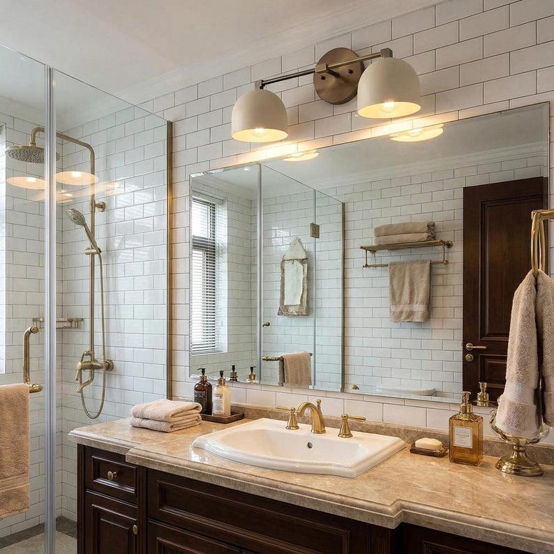 May include: A bathroom with a white tiled wall, a large mirror, and a dark brown vanity. The vanity has a marble countertop with a white sink and gold fixtures. A gold light fixture with two dome-shaped lights is mounted above the mirror.