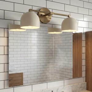 May include: A modern bathroom vanity light fixture with three off-white dome-shaped shades. The fixture has a brass finish and is mounted above a rectangular mirror. The background features white subway tile and wooden accents.
