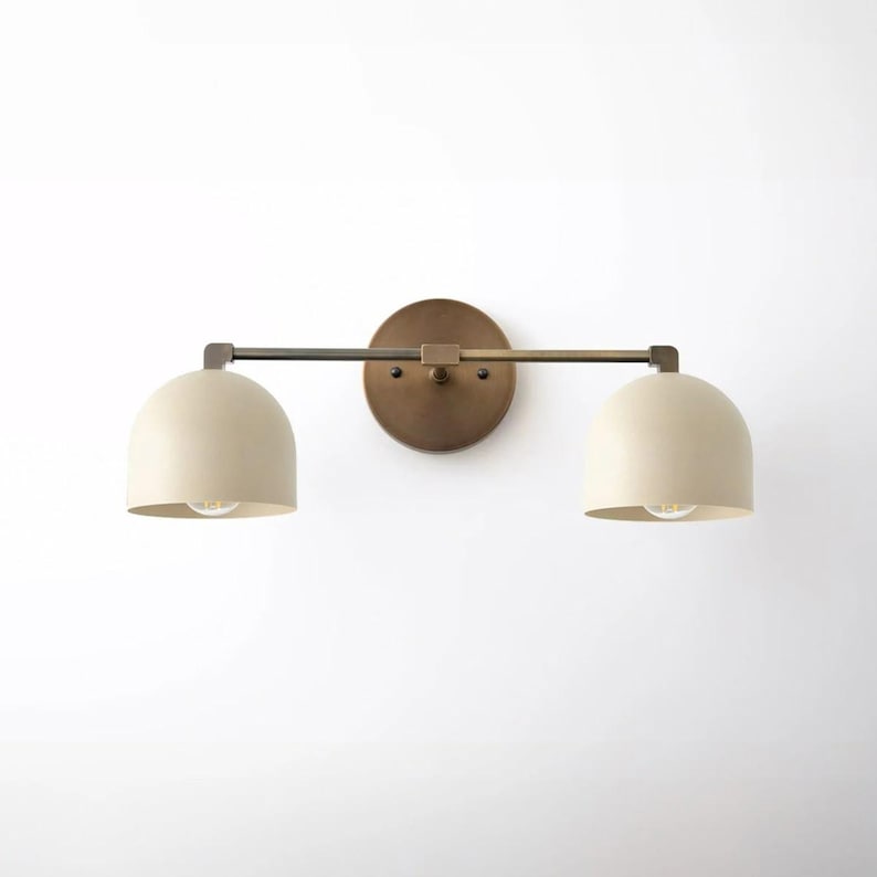 May include: A two-light wall sconce with a round, brass-colored backplate and a horizontal bar. Two off-white, dome-shaped shades are attached to the bar. The sconce has a modern design and is suitable for various interior styles.