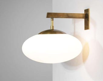 May include: A modern wall sconce with a brass finish. The fixture features a horizontal arm and a round, white glass shade. The sconce is mounted on a white wall, creating a minimalist aesthetic. The design is suitable for interior lighting.