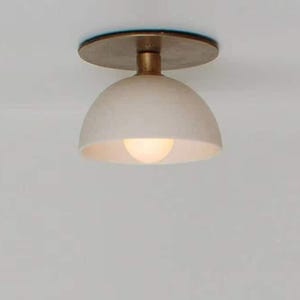 May include: A modern ceiling light fixture with a cream-colored, dome-shaped shade and a brass-colored stem and mounting plate. The light bulb is visible inside the shade, emitting a warm glow. The fixture is designed for indoor use.