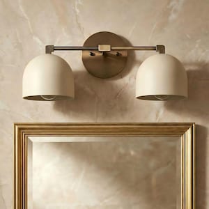 May include: A two-light vanity light fixture with a brass finish. The fixture has two off-white, dome-shaped shades and is mounted above a gold-framed mirror. The background is a marble-like wall.