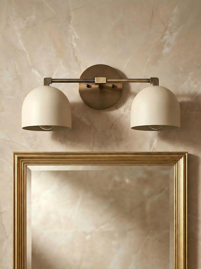 May include: A two-light vanity light fixture with a brass finish. The fixture has two off-white, dome-shaped shades and is mounted above a gold-framed mirror. The background is a marble-like wall.