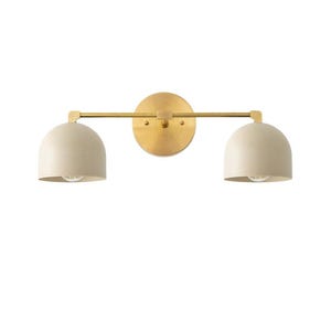 May include: A modern wall sconce with a brass finish. It features a circular backplate, a horizontal bar, and two cream-colored, dome-shaped shades. The sconce is designed for two light bulbs.