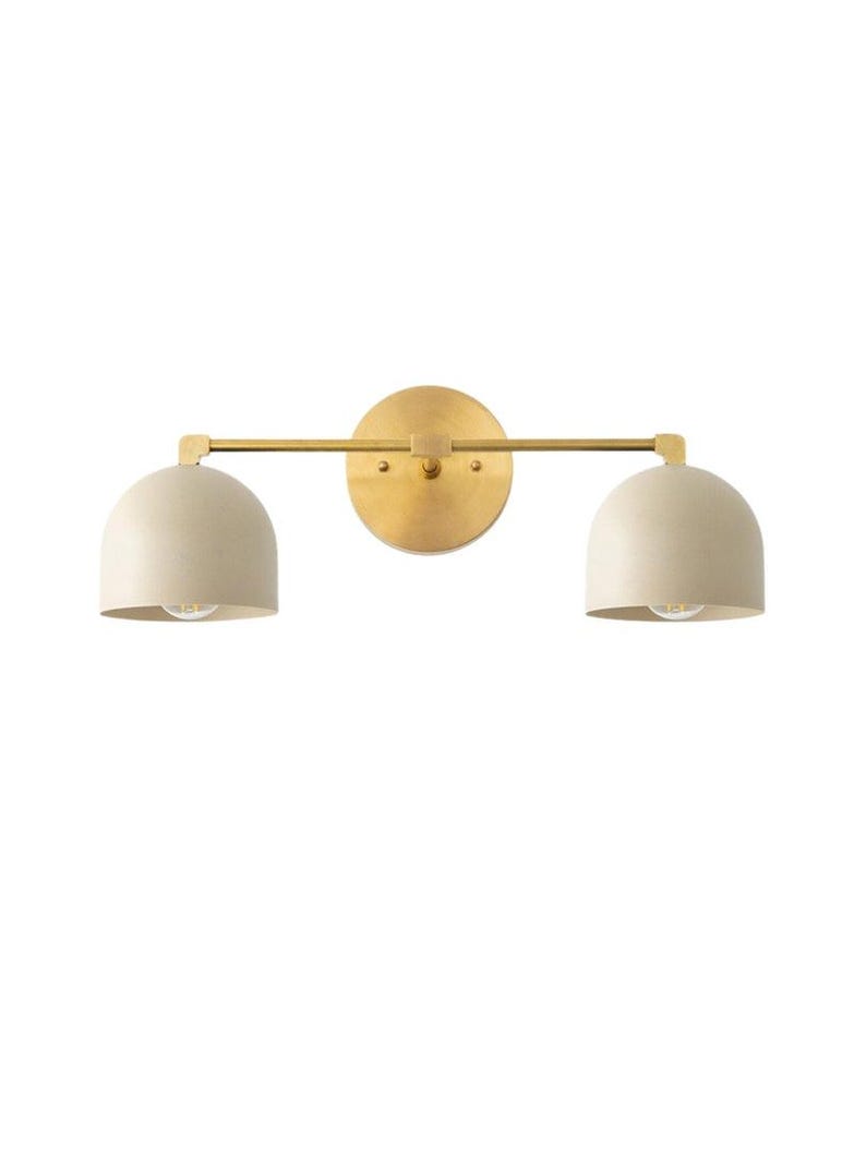 May include: A modern wall sconce with a brass finish. It features a circular backplate, a horizontal bar, and two cream-colored, dome-shaped shades. The sconce is designed for two light bulbs.
